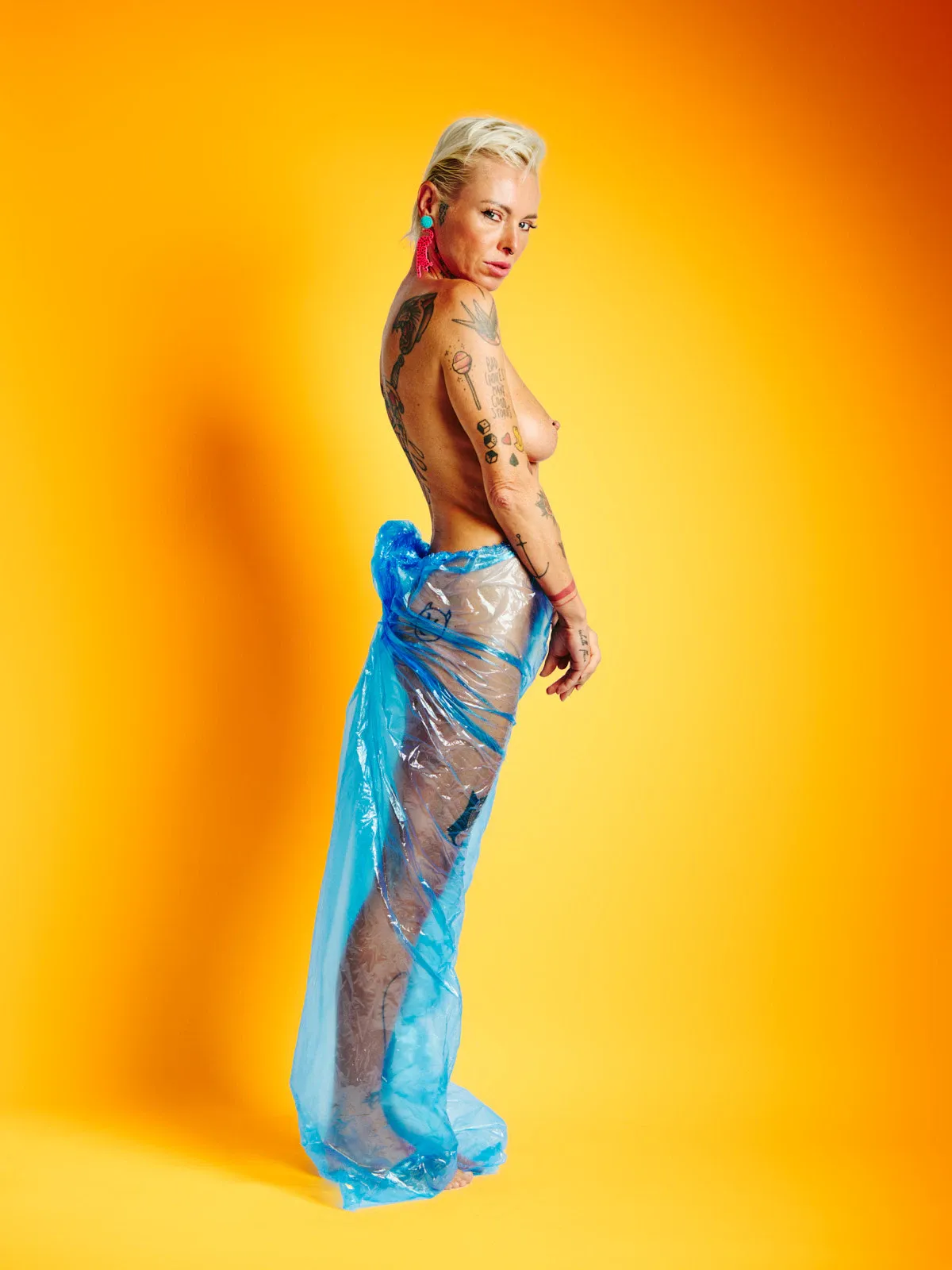 Lu Epic nude wearing blue transparent plastic pants against a strong yellow background in the Colors series