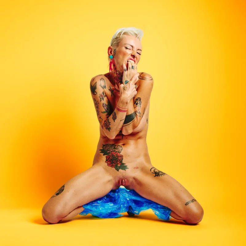 Explicit art nude portrait of Lu Epic in blue plastic pants with vivid yellow backdrop