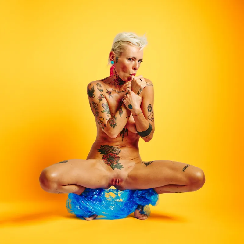 Lu Epic nude wearing blue transparent plastic pants against a strong yellow background in the Colors series