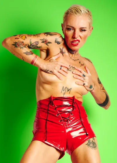 Lu Epic topless wearing red latex shorts and red high heels against a green background, expressing playful energy