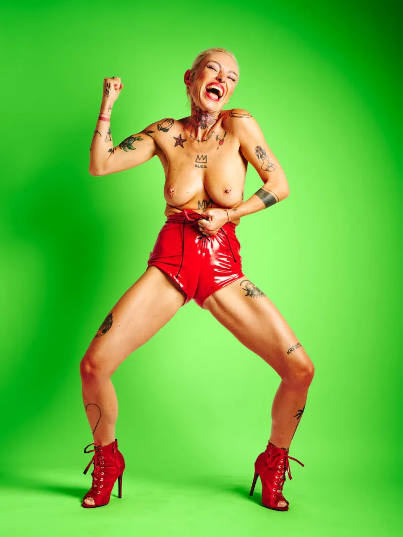 Explicit art portrait of Lu Epic topless in red latex shorts and high heels on a green backdrop
